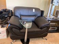Front view of black bonded leather chair with cushioned roll arms and chrome feet. The chair is placed on a carpeted floor with the tempered glass top side table partially visible.