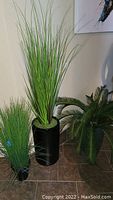Full view of three faux plants including two tall grass plants in large black pots and a smaller fern-style plant with detailed leaves.
