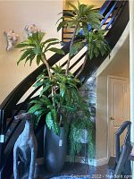 Full view of large faux tree with broad green leaves next to staircase and sitting on floor, in textured tall grey plastic planter, approx 31 inches tall pot.
