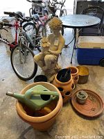 Garden fountain statue seated figure holding birdhouse, terra cotta pots varying sizes, plastic watering can, black plastic pot, and pot base tray visible in garage setting