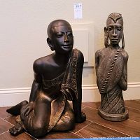 Two sculptures side by side against wall: bronze-tone kneeling figure and grey tribal bust.