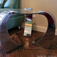 Side view of round glass table with prominent curved polished chrome metal legs showing design and structure.