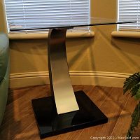 Side view of side table showing curved metal pedestal, black base, and clear glass rectangular top.