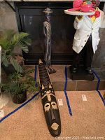 Photo showing the carved African wall mask lying on the floor alongside the human figure sculpture standing by the fireplace. The mask is long, black with beige and white carved face details.