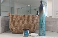 Photo shows basket, tall slender ceramic vase, small white soap dish, and eucalyptus tea candle arranged on countertop near mirror.