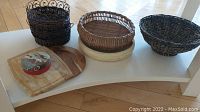 Overview photo showing all items including five woven baskets, wooden bowl, cutting board, storage tin, and wooden plate arranged together on a white table.