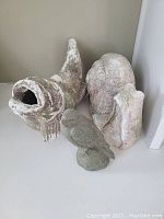 Three weathered outdoor cement statues: a fish with open mouth and curled tail, a snail with a large shell, and a small bird statue. Surfaces show moss and chipped paint.
