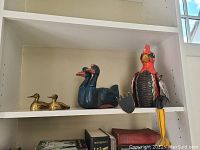 Four duck and chicken figurines displayed on a white shelf: two small brass ducks on the lower shelf; wooden two-headed duck and wooden minuet style chicken on the upper shelf.