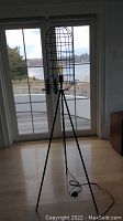 Full view of black industrial floor lamp standing on wood floor near window with outdoor view, showing tripod base and cage wire shade with visible bulb inside, power cord laid out on floor.