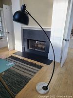 Full view of the black metal curved floor lamp standing on a wood floor with a white marble base visible at the bottom. Lamp bends outward with shade facing downwards.