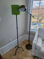 Two metal floor lamps, one with green shade and tripod base, other adjustable with dome shade, placed near window and couch.