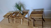 Four IKEA wooden folding slatted tables/stands with varying wood tones, one missing a top slat, placed next to each other with a faux grass plant in a glass vase on top of one table.
