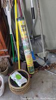 Multiple garden tools with long handles leaning against a wall including broom, rake, hoe, and a yellow roll of weed block cloth standing upright.