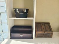 Three office organizers shown in shelving: one rectangular metal black bin with silver handle and rivet details on top shelf, two wood organizers below, one with compartments and small drawers