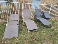 Four patio lounge chairs placed outside near a fence by dried plants and grass with a body of water visible behind the fence.
