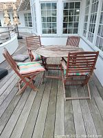 Wide view of octagonal teak patio table surrounded by four teak chairs on outdoor deck with cushions on seats showing weathering and use wear.