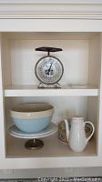 Full view of kitchen shelf with all items: ceramic bowl, cake stand, covered dish, coffee pot and vintage scale