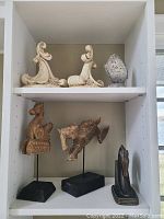 Photo showing two white wave-shaped bookends with scroll designs on top shelf and lower shelf holding two wood horse sculptures, one missing tail, and a silver decorative piece