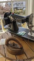 Photo shows black antique Singer sewing machine with electric motor, and cast iron antique iron with wooden handle on wooden surface by window.