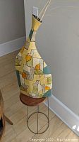 Tall decorative vase on metal base stand with wooden top, geometric multicolor design.