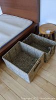 Two woven storage baskets and one wooden stool placed on a hard floor near a bed frame.