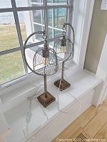 Both matching lamps on a white window sill showing full height and design with chicken wire cages and wooden bases.