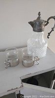 Vintage glass pitcher with ornate silver handle and spout, two glass holders with silver-ringed holders on a painted wooden surface.