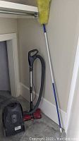 Full view of Kenmore canister vacuum cleaner standing with attached hose and motorized brush, yellow duster with blue handle, and plastic hangers leaning against wall.