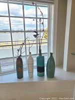 Set of four tall ceramic vases on a window ledge with a view outside. Three of the vases contain dried branches.