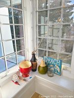Two wood vases, glass vase with branches, lidded red urn, and a blue Beach sign reproduced art piece on a window sill