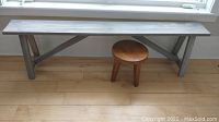 Front view of wooden long coastal style bench next to round wooden stool on light hardwood floor.