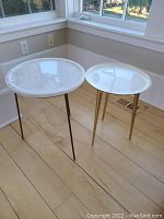 Two side tables placed near window on wooden floor; one with white marble lazy Susan top and dark metal tripod legs, the other a white round top with gold-toned tapered legs