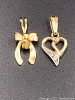 Two gold pendant charms on black background. Left is a bow design with a small stone, right is an open heart design with three small stones.