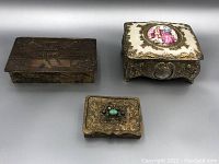 Three vintage decorative boxes displayed on a plain white surface: a rectangular engraved metal box, a small square metal box with turquoise stone, and an ornate metal box with porcelain top.