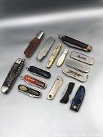 Overhead view of 14 vintage folding pocket knives and metal sheaths laid out on a gray background