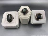Photo of three labradorite rings displayed on white ring stands: oval, teardrop, and square stones with silver bezels.