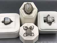 Four rings displayed on three white ring stands, showing the variety of moonstone shapes and silver decorative settings.