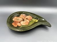 Top and angled view showing the oval curved spoon dish with a raised floral design featuring a peach-orange flower with green leaves on an olive green background, glossy finish.