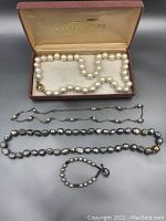 Overview of all 4 pieces including the white pearl necklace in the box and the gray baroque pearl necklace, silver chain necklace, and pearl bracelet laid out on surface.