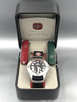 Swiss Military wristwatch in a Victorinox genuine Swiss Army knife branded box alongside a red and a green Swiss multi-tool knife.