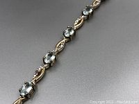 Close-up of section of the tennis bracelet showing round aquamarine-colored topaz stones and gold-tone metal links with intricate bead-like detailing, highlighting the prong settings and metal finish.