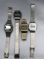 Photo showing four vintage digital wristwatches with metal stretch bands lying flat, from top to bottom: one silver-tone watch with black face, one Phasar perma-glow watch, one gold-tone watch, and one Artron watch, all in used condition
