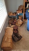 Multiple wicker and woven baskets arranged on carpet floor against wall including large rectangular basket with handle, small round and oval baskets, dark brown baskets, and picnic baskets.