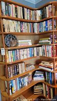 Wide view of wooden corner bookshelves filled with multiple rows of books of various sizes and colors with a black wall clock and some stacked books.