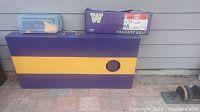 Photo of purple and yellow wooden cornhole board with hole, plastic container and golf set case below.
