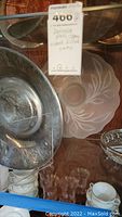 Photo showing an aluminum lazy Susan with an etched glass plate with floral designs inside alongside other glassware beneath glass shelf.