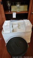 One photo showing white padded carrying case holding multiple glasses with white styrofoam compartments and black cast iron Dutch oven with lid in front.