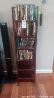Full view of tall solid wood DVD rack shelf unit holding DVDs and Blu-rays, standing on floor next to wall and speakers.