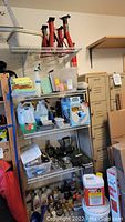 Full shelf view showing the jack stands, plastic tubs, towels, assorted spray bottles, automotive fluids, tools, and cleaning supplies organized on white wire shelving.