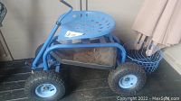 Full view of blue metal rolling garden chair with perforated seat, crank handle, and attached wire basket behind the seat.